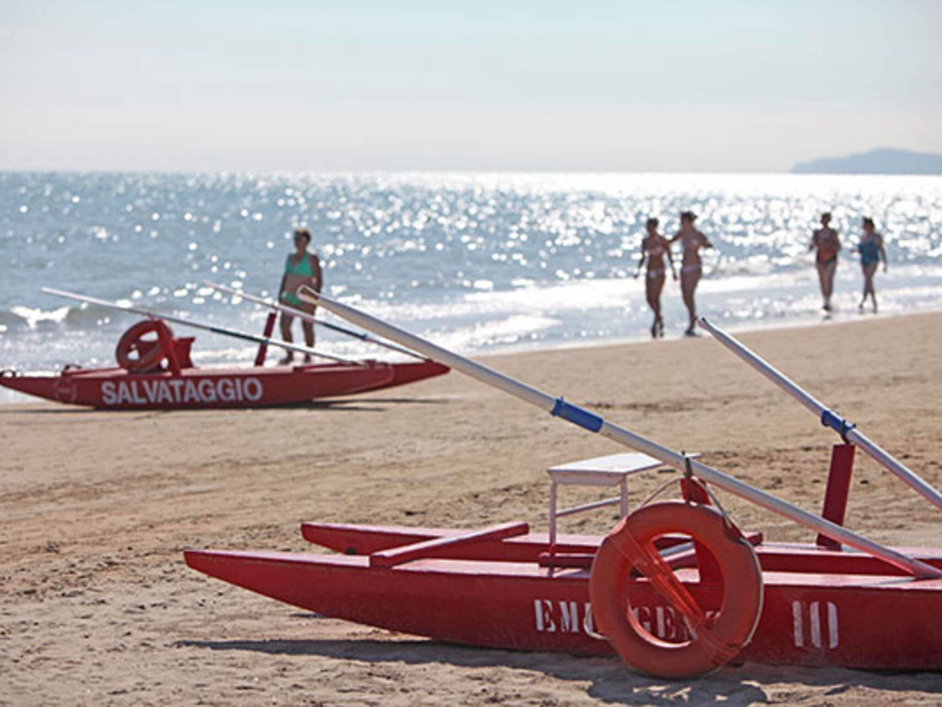 Un bote rojo con la leyenda 'SALVATAGGIO' y 'EMERGENCIA' escrito, sobre una playa arenosa con el océano al fondo.