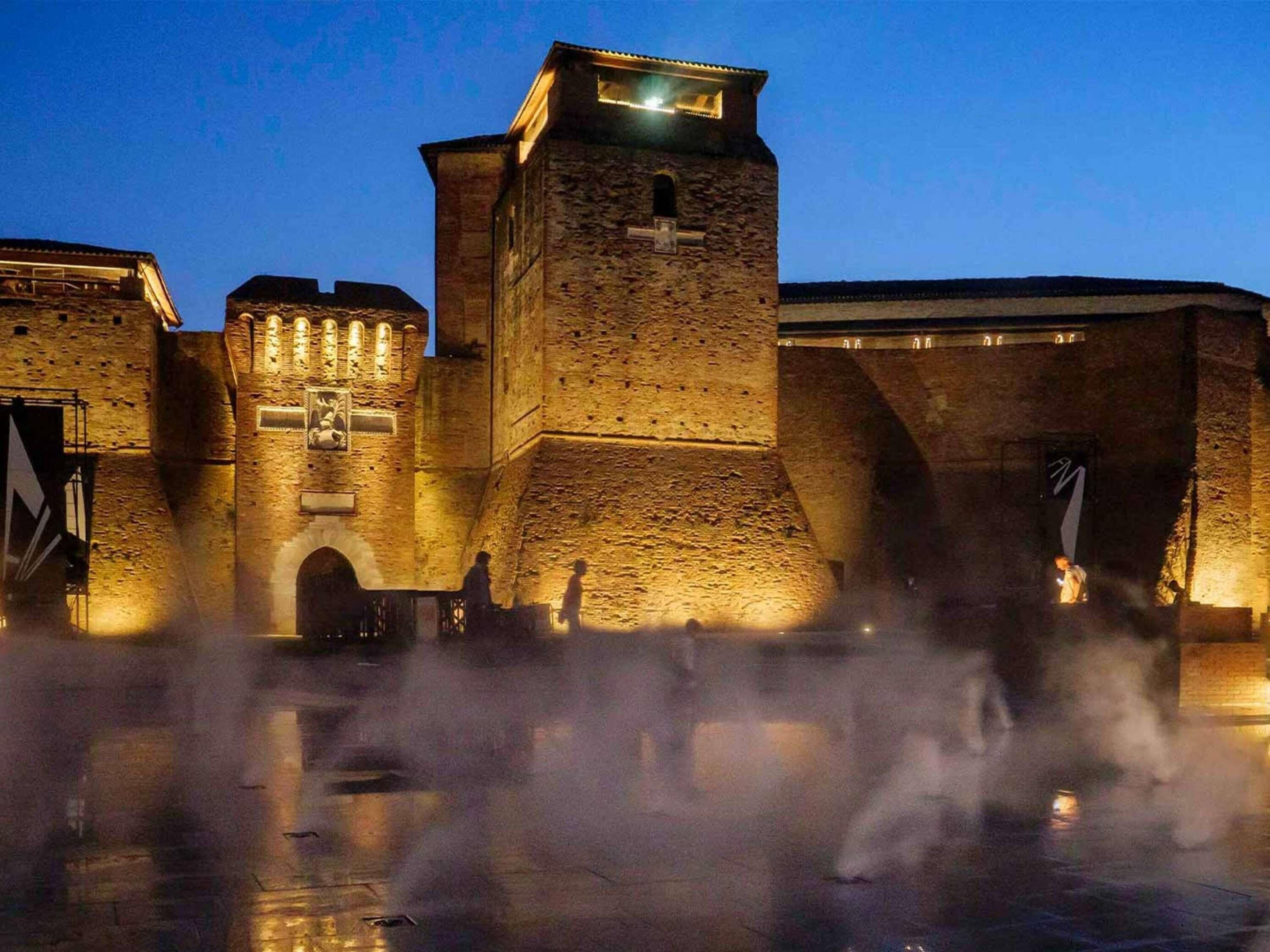 A historic building with a prominent tower, arched gateways, and a reflective water feature.
