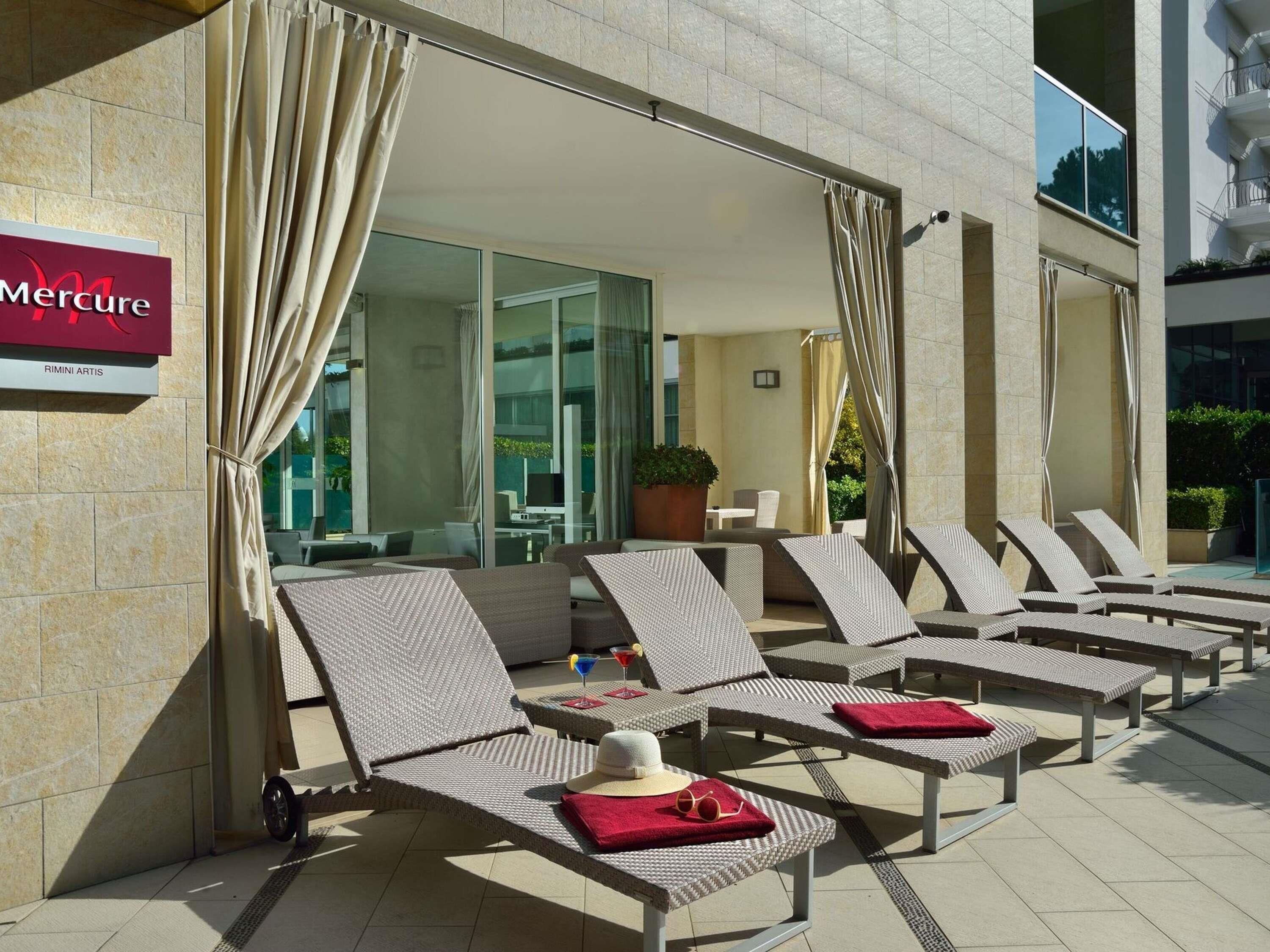 A hotel exterior with a lounge area featuring sun loungers, a sign for 'Mercure,' and a glass door entrance.