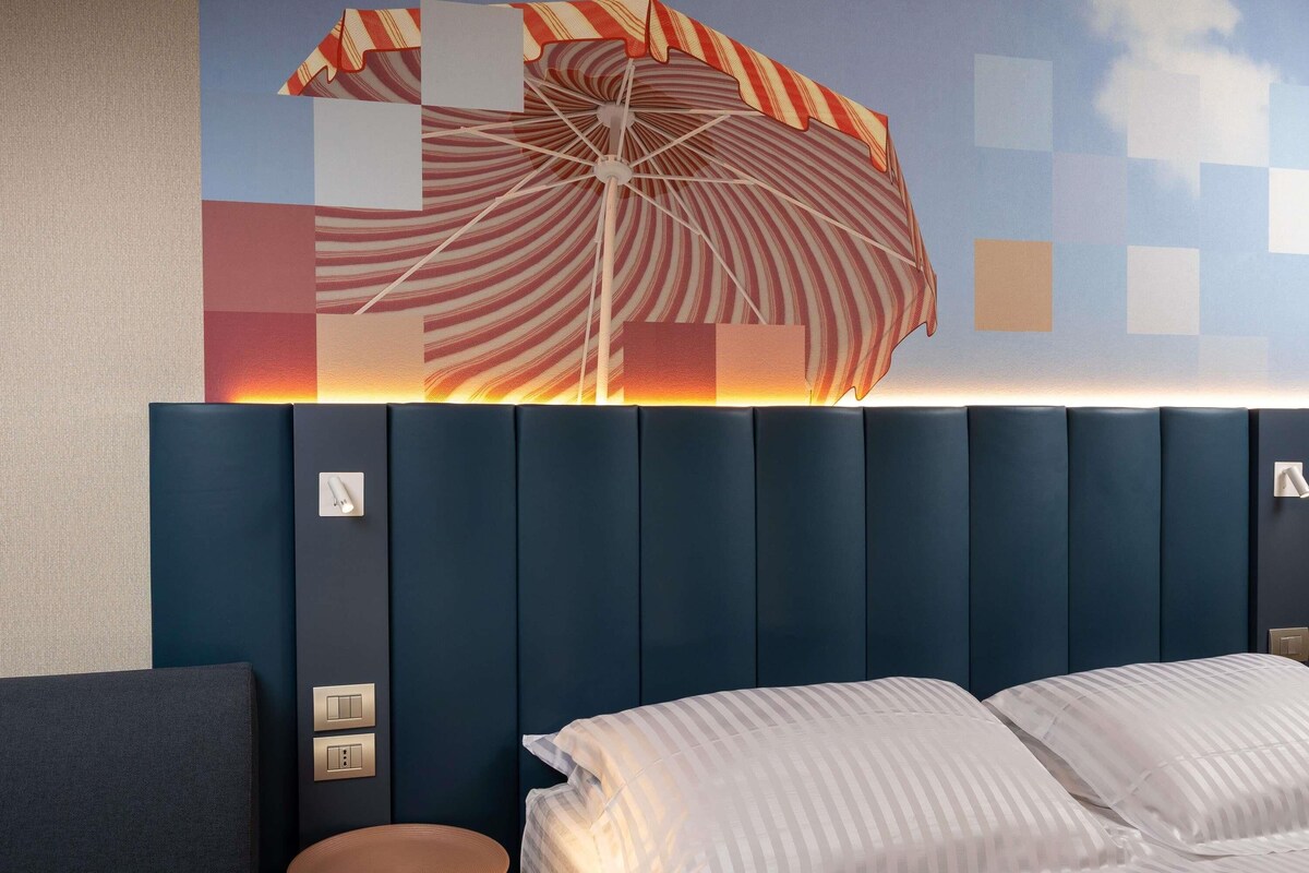 Quarto de hotel com cama, mesa redonda pequena e luminária fixada na parede.