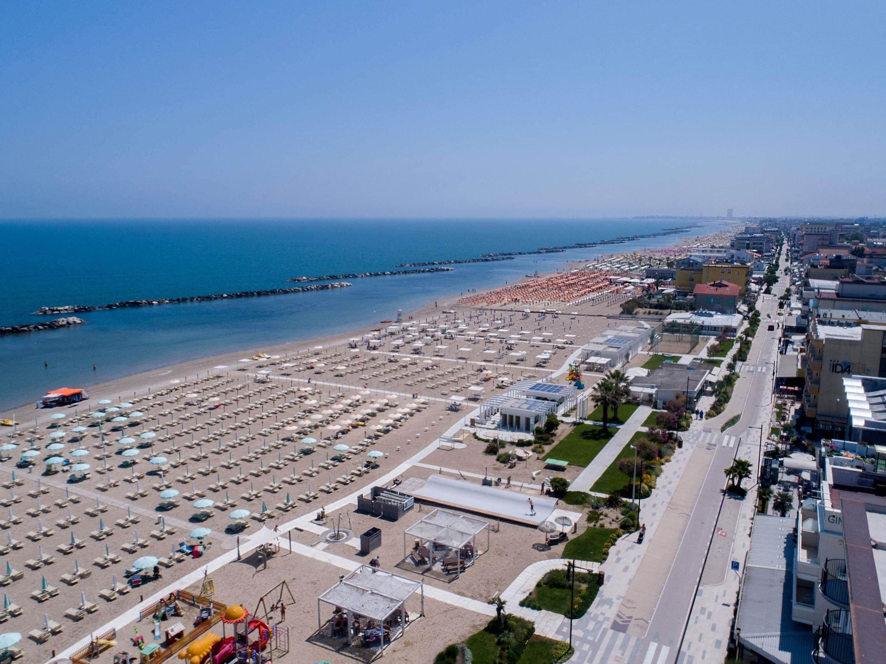 An aerial view of a beach with rows of sunbeds and umbrellas, a promenade, and buildings along the coastline.