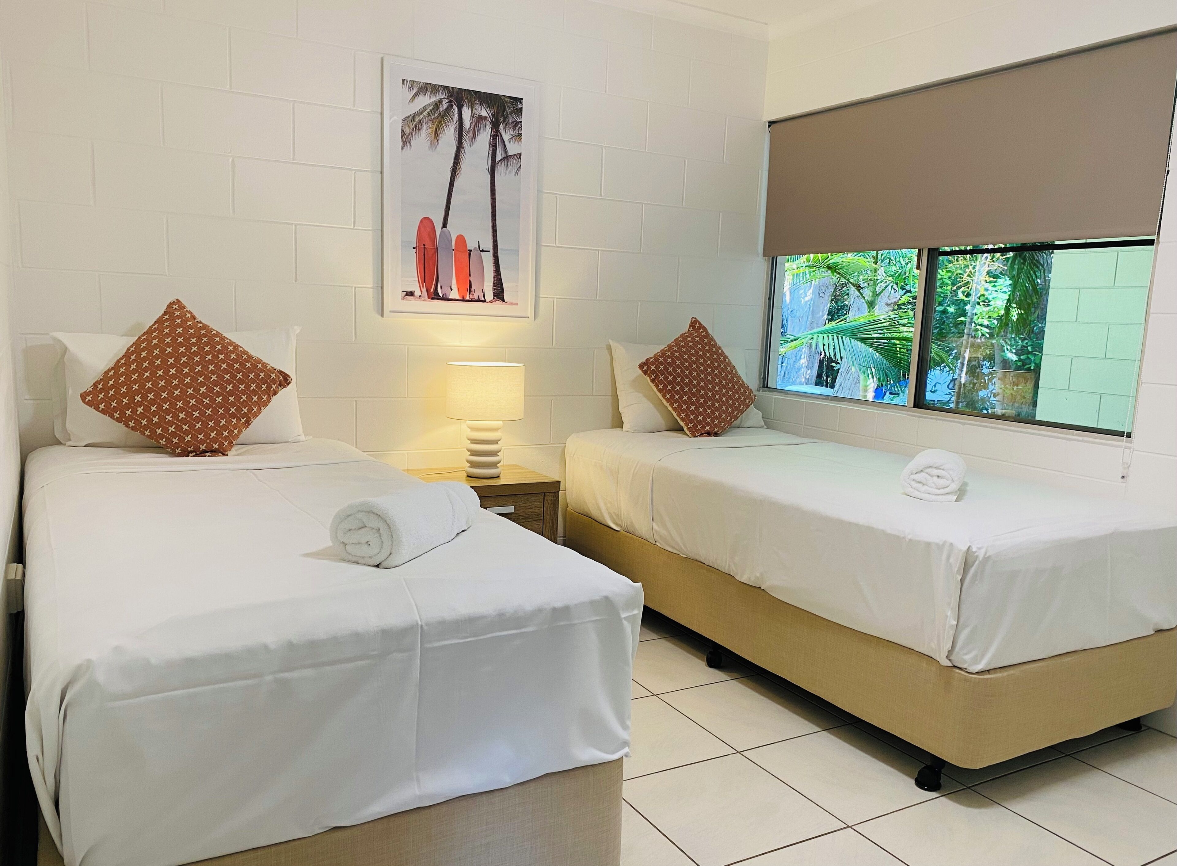 Camera d'albergo con due letti, biancheria da letto bianca, cuscini marroni a motivi decorativi, un comodino con una lampada, un quadro appeso al muro e una finestra con vista sul verde.