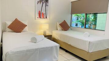 Camera d'albergo con due letti, biancheria da letto bianca, cuscini marroni a motivi decorativi, un comodino con una lampada, un quadro appeso al muro e una finestra con vista sul verde.