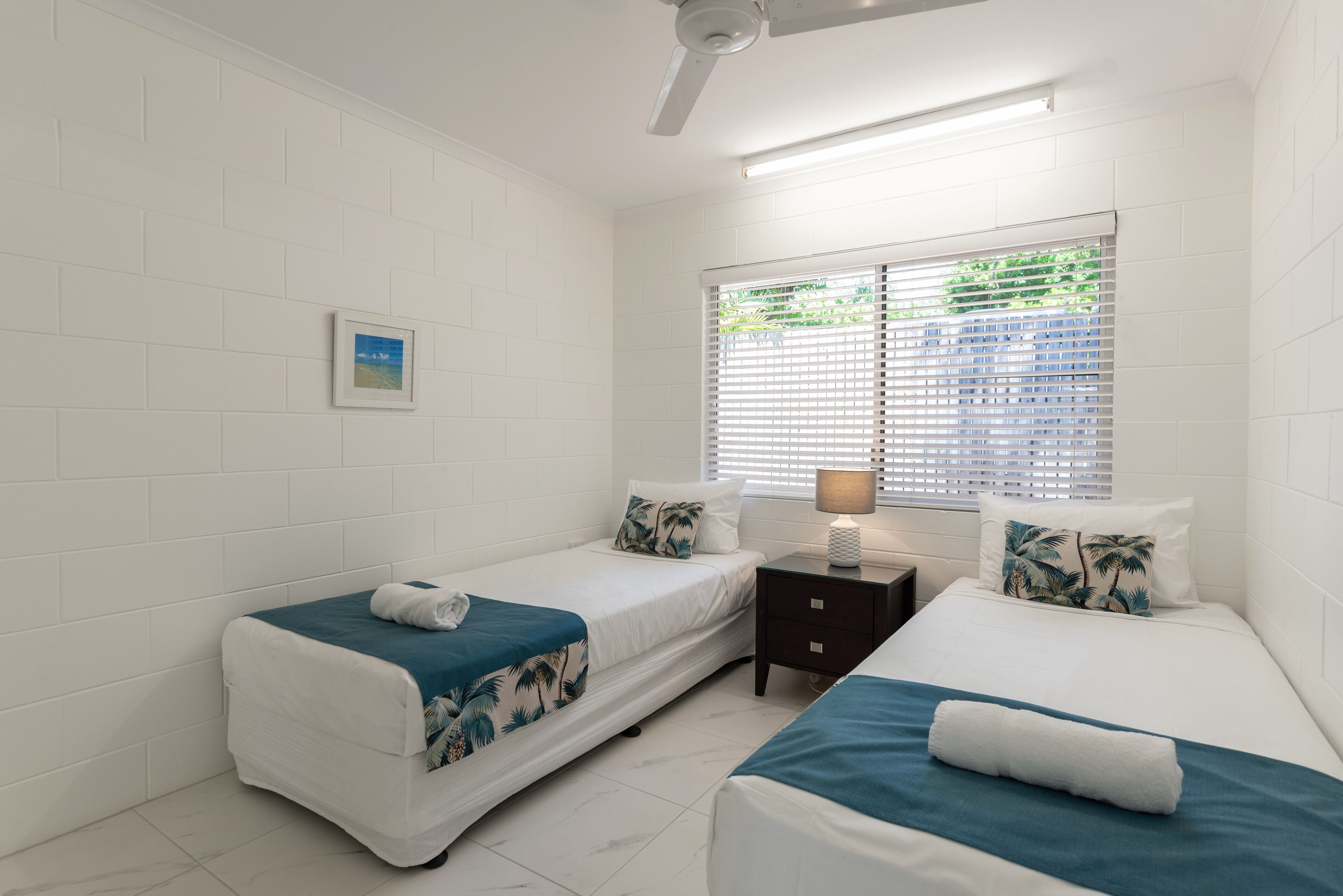 A room with two beds, white walls, a ceiling fan, a window with blinds, a small table with a lamp, and a framed picture on the wall.