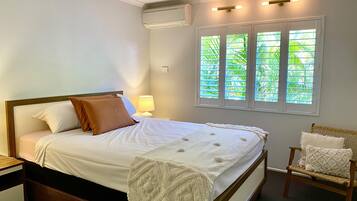 Un lit bien fait, avec une couette blanche et des oreillers marron, une lampe de chevet, une table de chevet en bois, une chaise en osier avec un coussin, et une fenêtre avec des stores donnant sur la verdure.