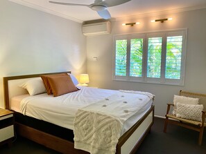 Una cama bien hecha con una colcha blanca y almohadas marrones, una lámpara de noche, una mesita de madera, un sillón de mimbre con cojín y una ventana con persianas que da a vistas de follaje.