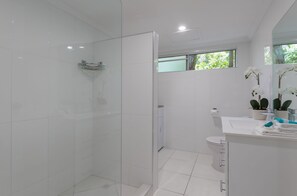 Un baño moderno con azulejos blancos, cerramiento de ducha de vidrio, inodoro, lavamanos con espejo y ventana con vistas a zonas verdes.