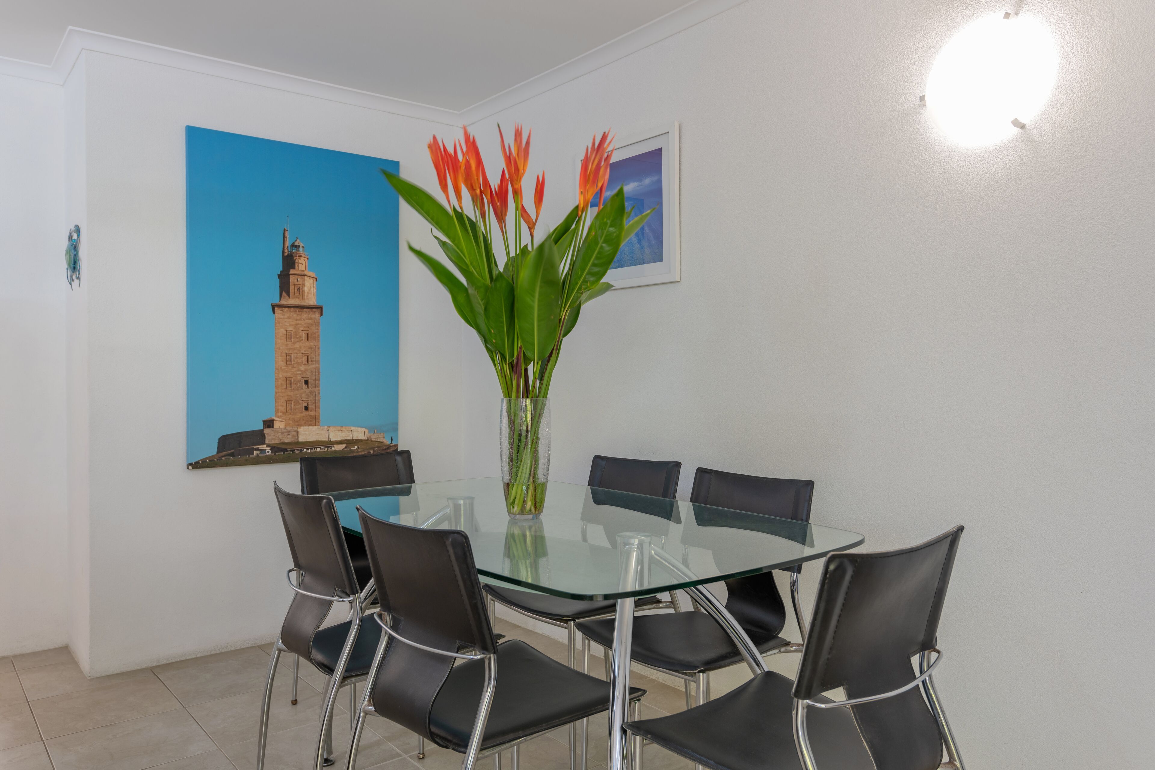 A modern dining area with a glass table and black chairs, a decorative vase with flowers, and a framed picture of a tower on the wall.