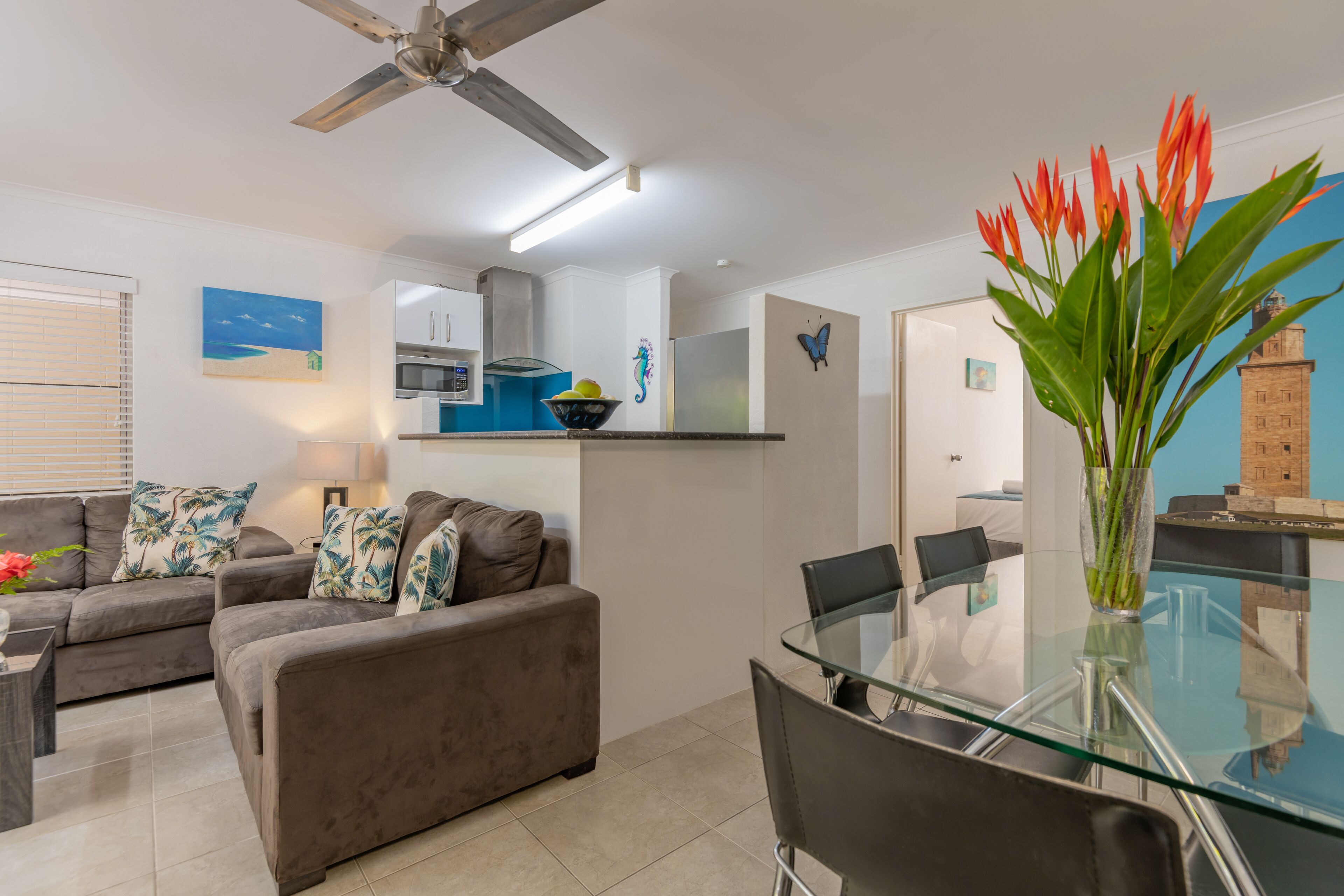 A modern living room with a glass dining table, two sofas with tropical cushions, a ceiling fan, and a kitchen area in the background.