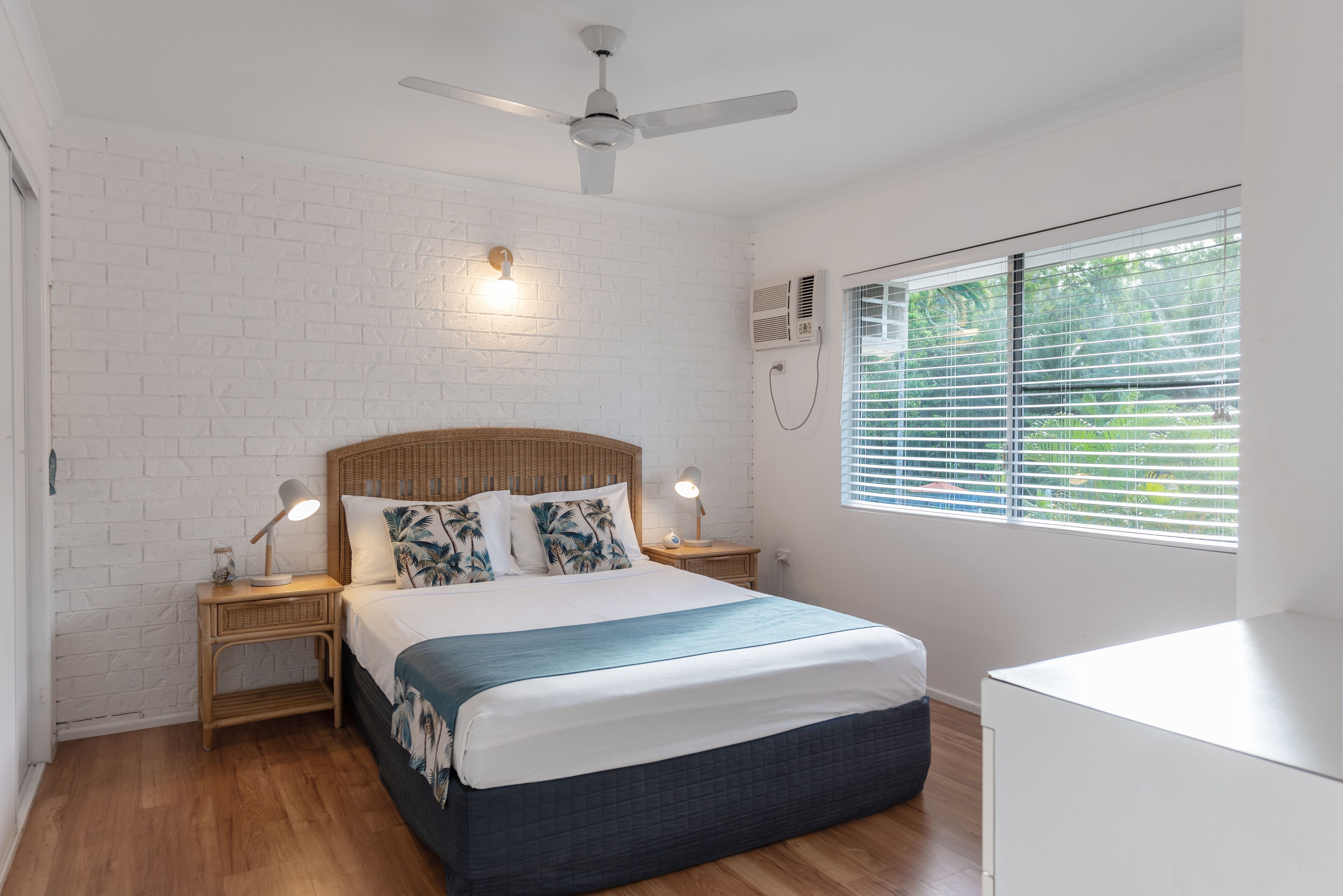 Quarto com parede de tijolos brancos, ventilador de teto, cama com colcha azul e branca, duas luminárias de cabeceira e uma janela com persianas.