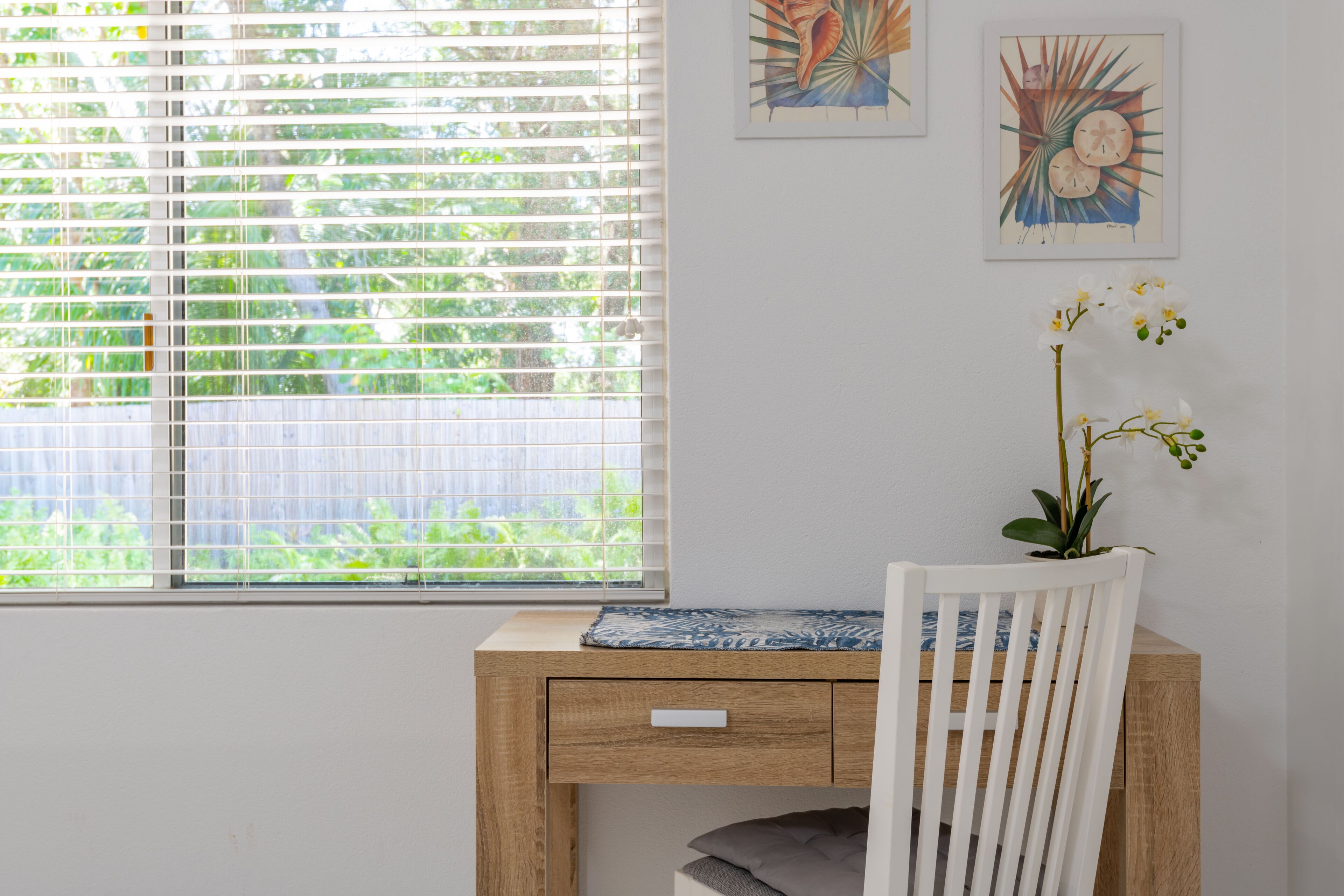 A room with a wooden desk, a white chair, a window with blinds, and two framed artworks on the wall.