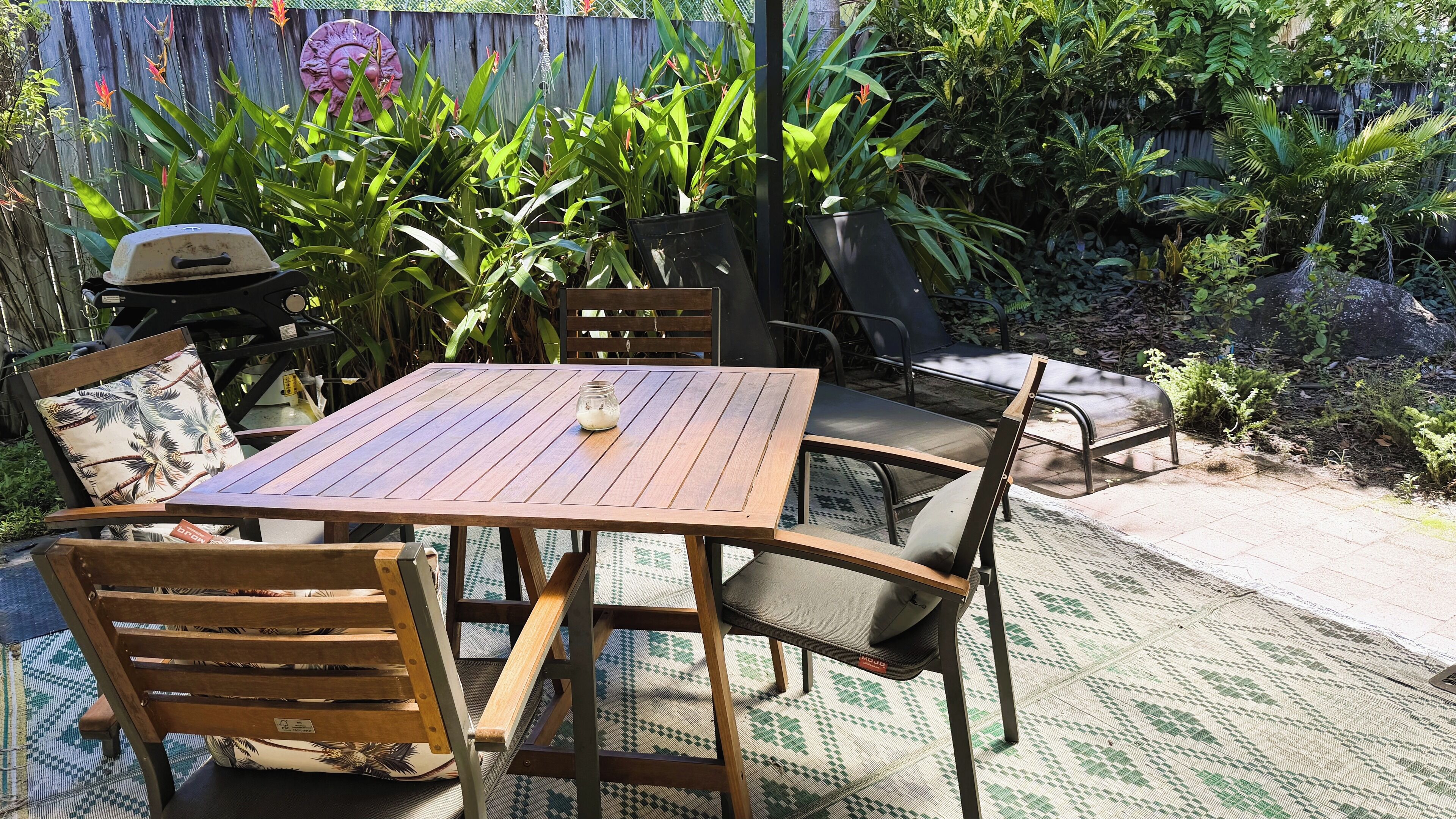 Une terrasse extérieure avec une table et des chaises en bois, entourée d’une végétation luxuriante et d’une clôture en bois.