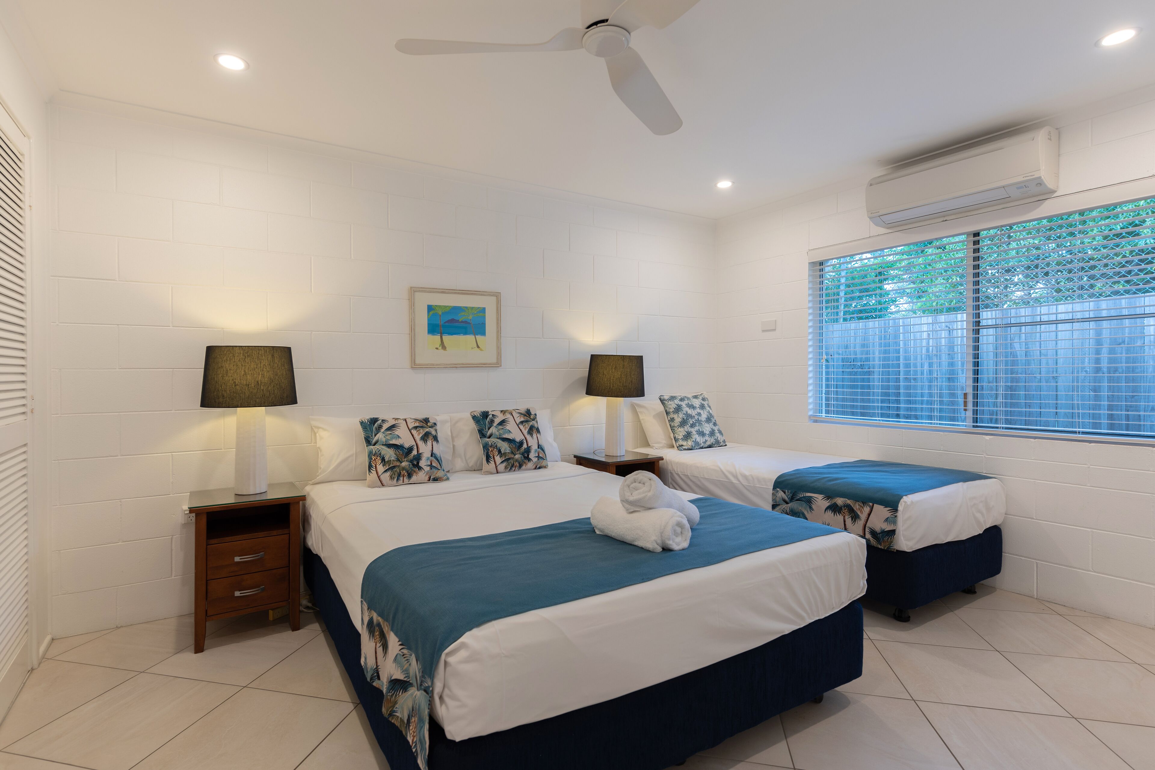 Habitación de hotel con dos camas, ventilador de techo, equipo de aire acondicionado y ventana con persianas.