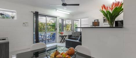 Une cuisine moderne avec un coin repas, dotée d’un ventilateur de plafond, d’un saladier de fruits sur la table et offrant une vue sur l’extérieur.