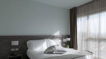 Habitació d'hotel amb un llit gran, una taula de nit amb una làmpada i una finestra amb cortines translúcides.