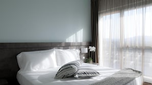 Una cama bien tendida con sábanas blancas y un plaid gris, una mesita de noche con lámpara, y cortinas traslúcidas que dejan entrar luz natural.