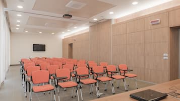 Un salón de conferencias con filas de sillas naranjas, una mesa de madera con una computadora portátil y un proyector en el techo.