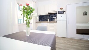 A compact kitchen with white cabinets, a microwave, and a refrigerator. There's a dining table with a patterned tablecloth and a vase with flowers. - Divine Apartments (Dubrovnik)