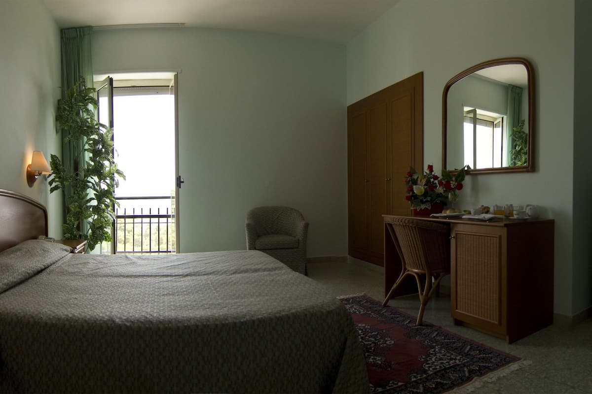 A hotel room with a bed, bedside table, lamp, chair, desk, mirror, dresser, television, and a window with curtains.