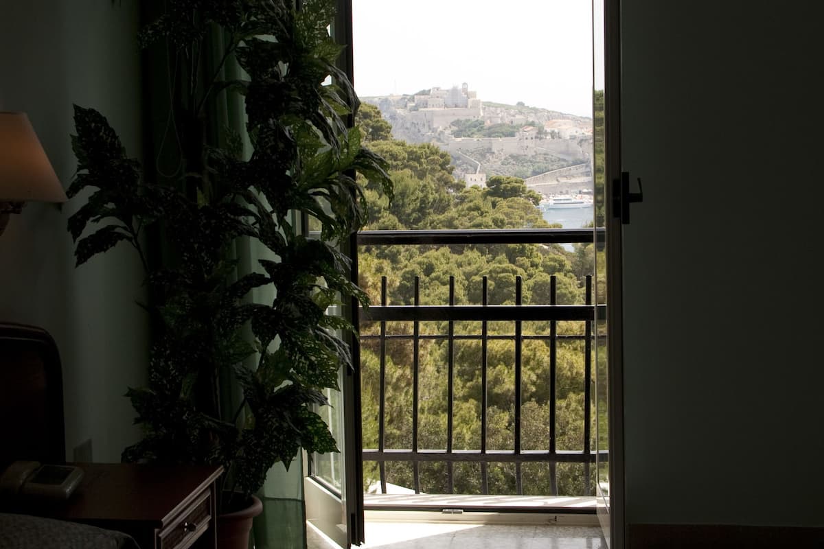 A hotel room with a bed, a desk, a potted plant, and a balcony with a view of a cityscape.