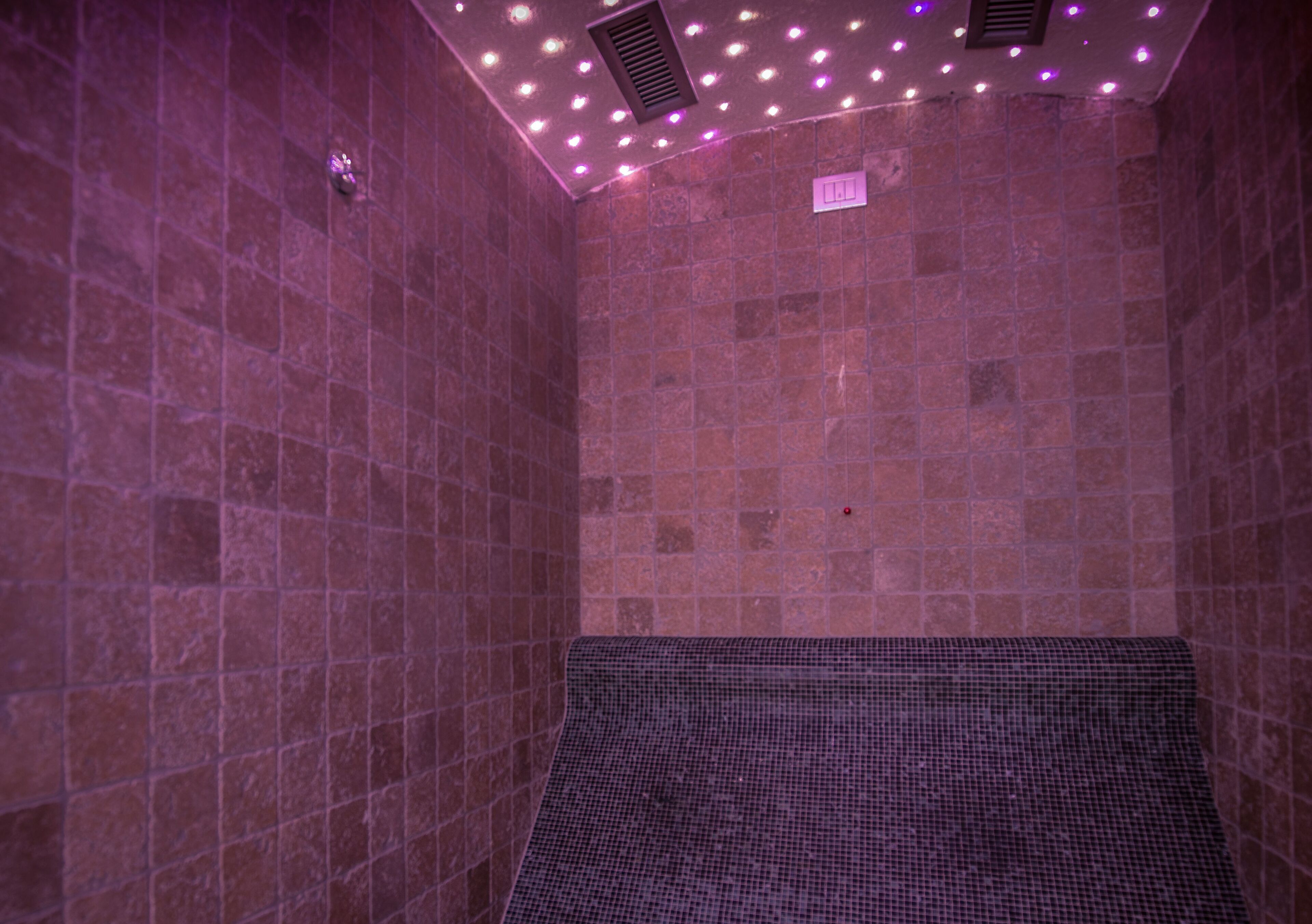 A room with tiled walls and a textured floor, equipped with a ceiling vent and a small sign.