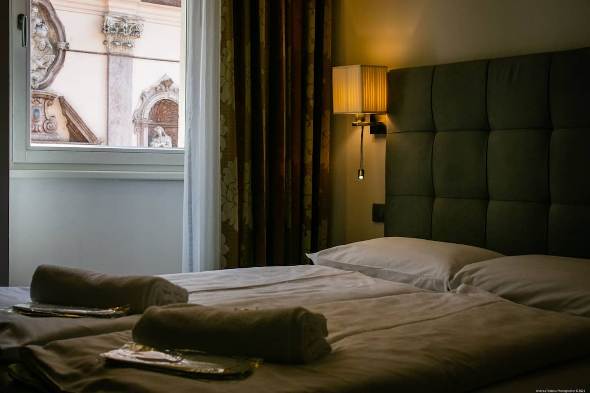 A hotel room with two beds, a window with curtains, a wall-mounted lamp, and a decorative mirror.