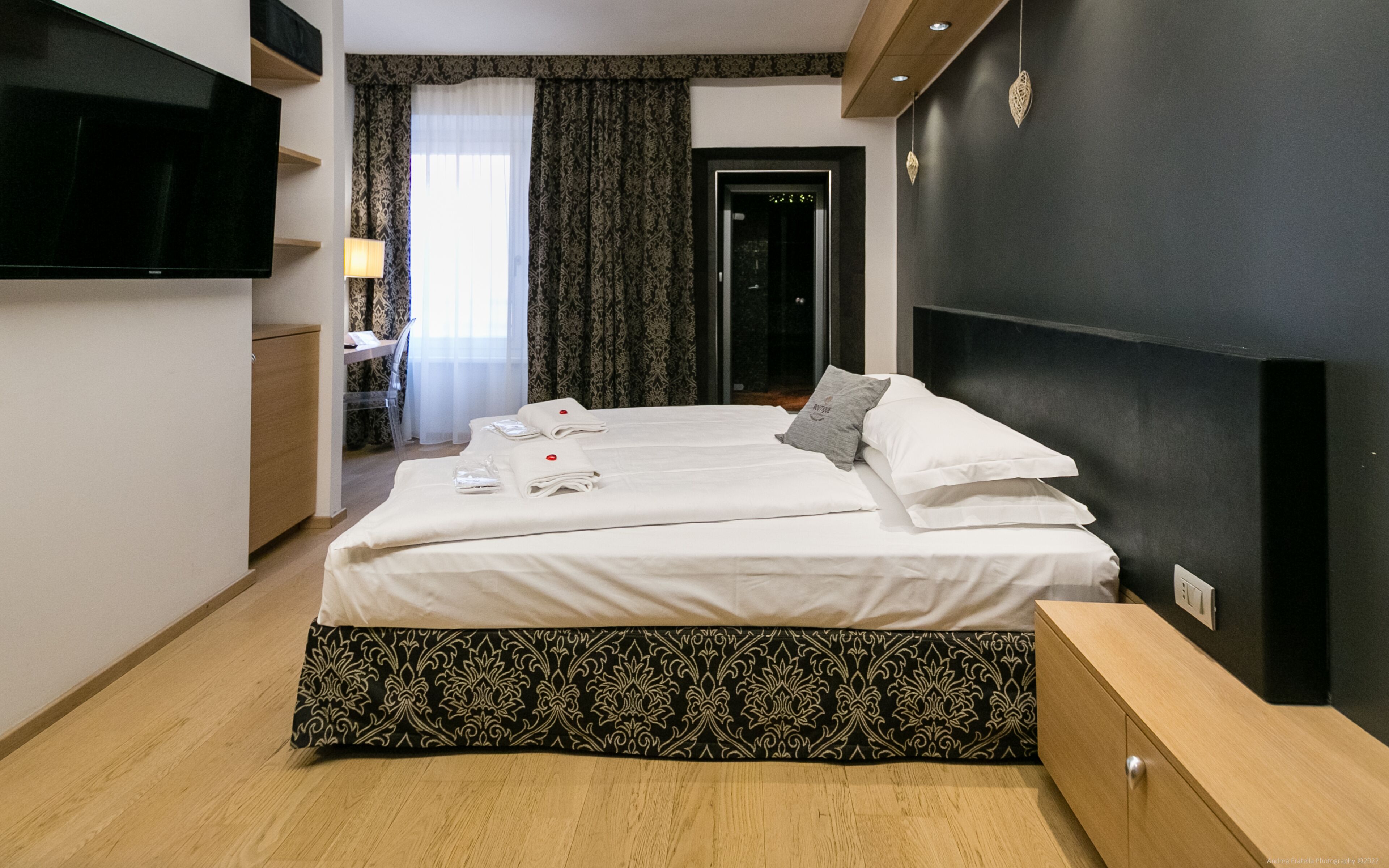 A hotel room with a bed, bedside tables, a TV, and a window with curtains.