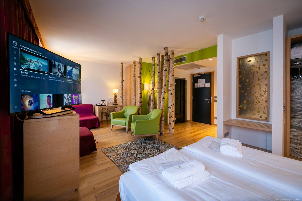 A hotel room with a bed, a desk with a flat-screen TV, a green armchair, a red sofa, and a patterned rug.