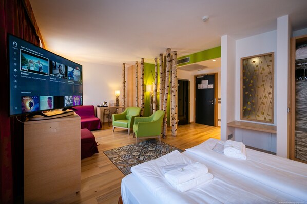 A hotel room with a bed, a desk with a flat-screen TV, a green armchair, a red sofa, and a patterned rug.