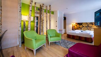 Moderni hotellihuone, jossa on kaksi vihreää nojatuolia, puulattia, sängynpääty, jossa on puunkuvio, ja suuri ikkuna.