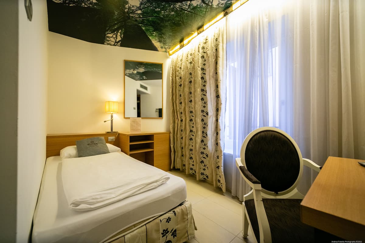 A hotel room with a bed, a desk, a chair, a lamp, a mirror, and curtains.