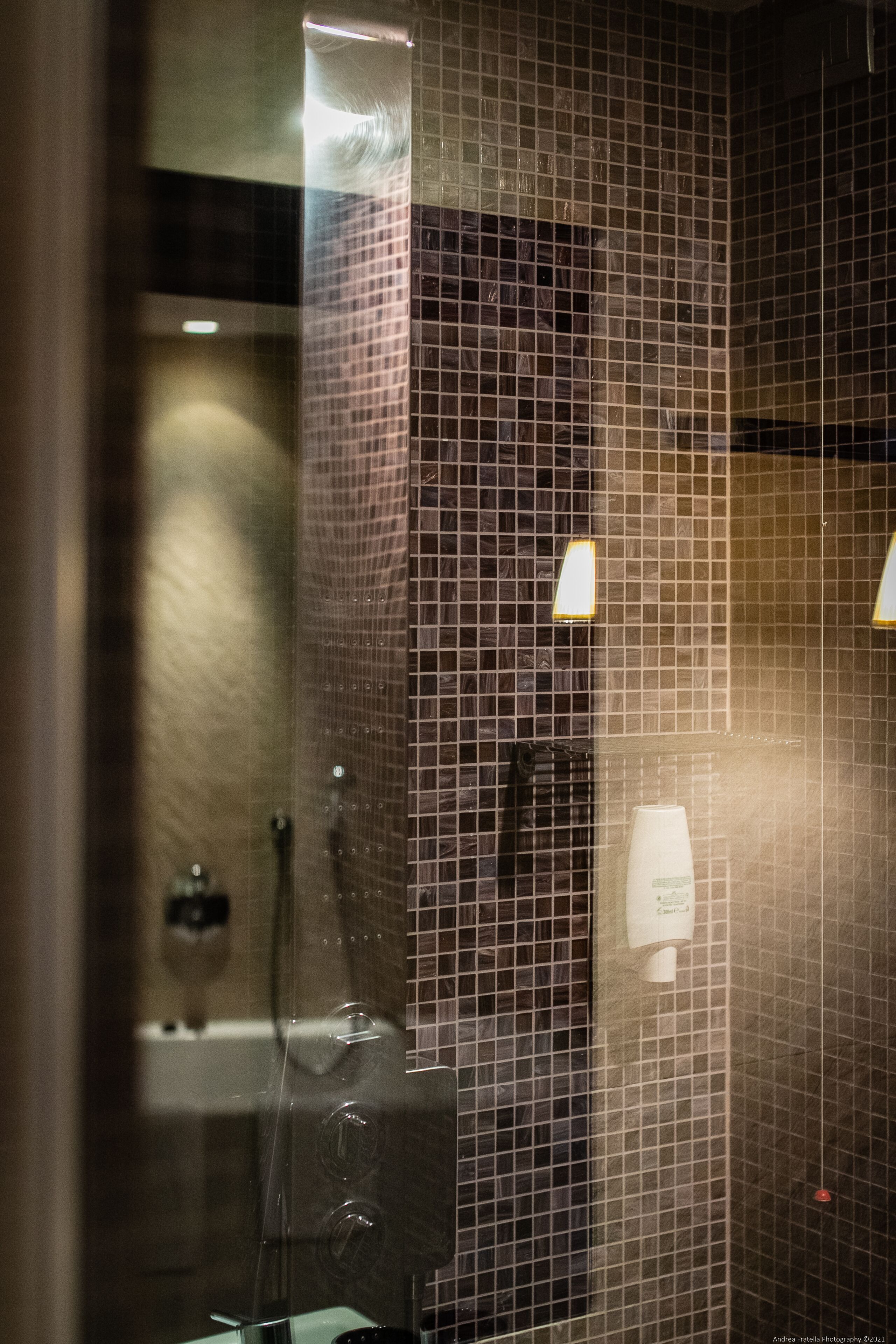 A bathroom with a tiled wall, a glass shower enclosure, and a white soap dispenser.