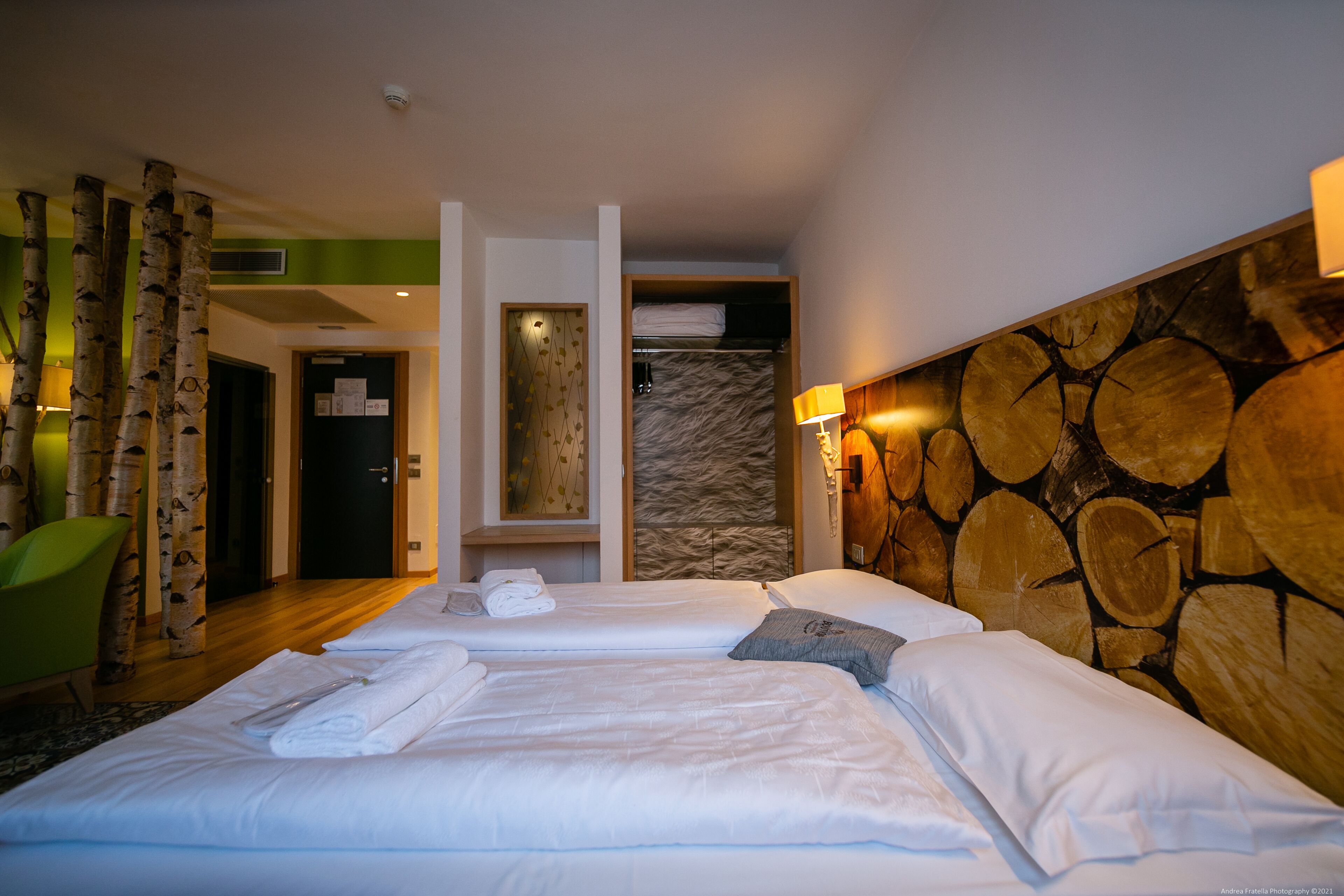 Une chambre d’hôtel avec deux lits, un fauteuil vert, une armoire et un mur décoré d’un motif imitant des troncs d’arbres.