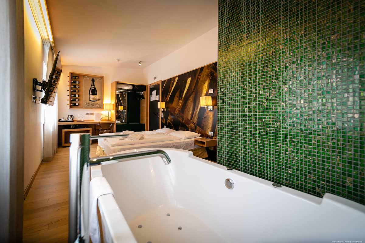 A modern hotel room with a large bathtub, a wooden floor, a green mosaic wall, and a wine rack with bottles.