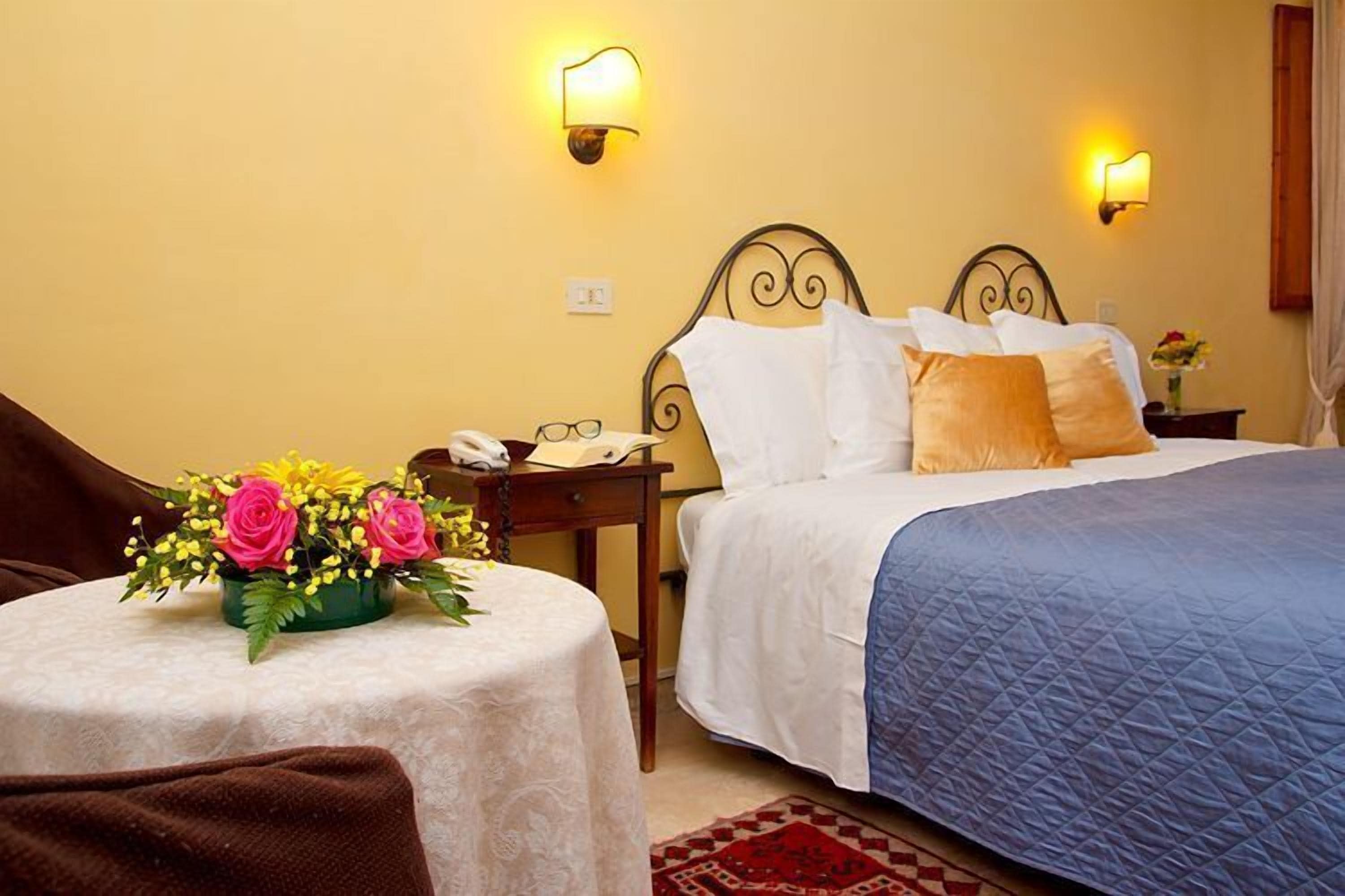 A hotel room with a bed, bedside tables, a small table with a floral arrangement, and wall-mounted lights.