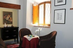 A room with a wooden ceiling, a black dresser, a red table with a vase of flowers, and a window with a wooden frame and yellow curtains. - Albergo Duomo (Montepulciano)