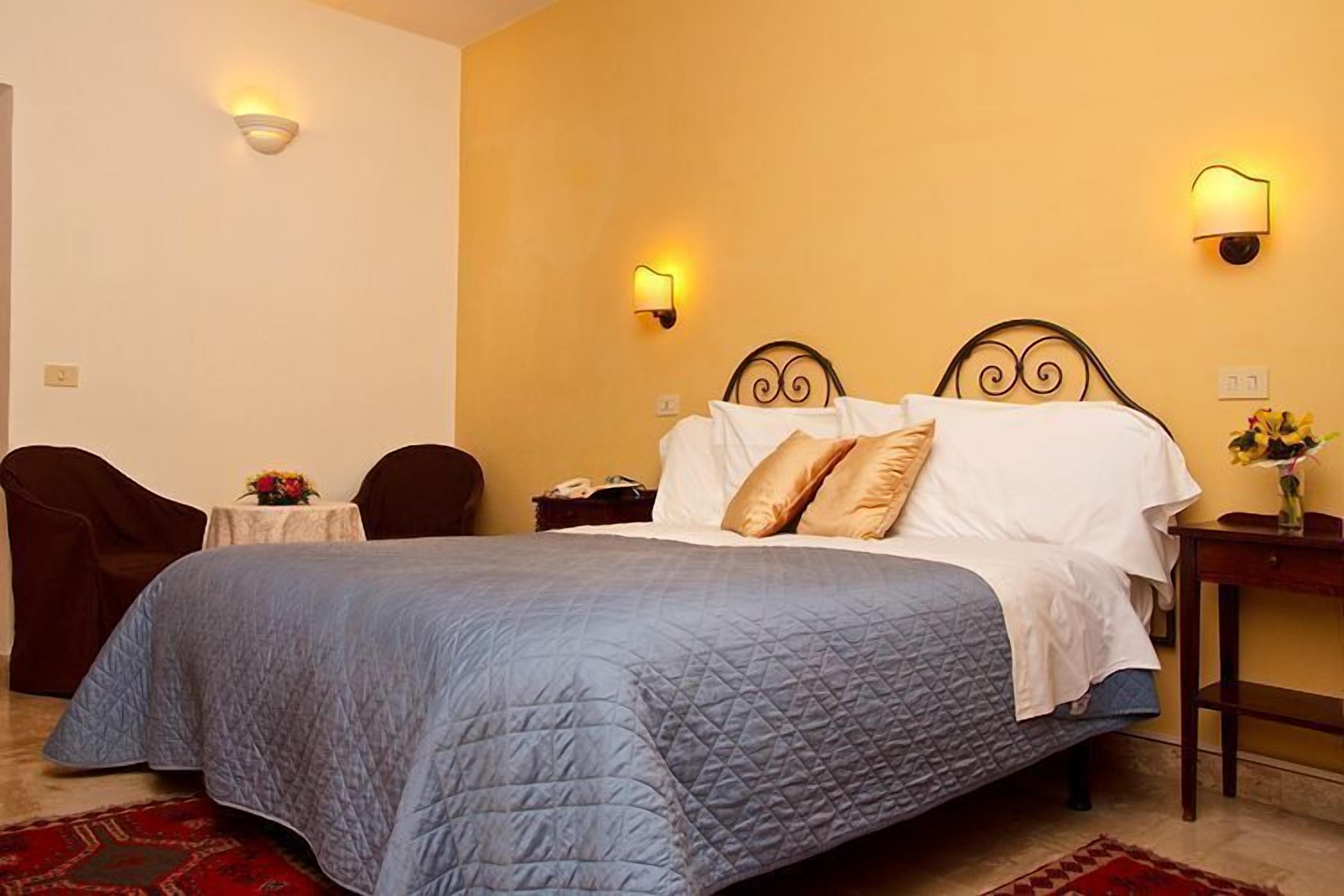 Une chambre d’hôtel avec un grand lit, deux lampes fixées au mur, une petite table sur laquelle est posé un vase de fleurs et un tapis rouge à motifs.