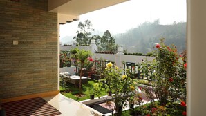 A balcony with a stone wall, a view of a garden with various plants and flowers, and a distant landscape with trees and hills. - Elysium Gardens Hill Resorts (Devikolam)