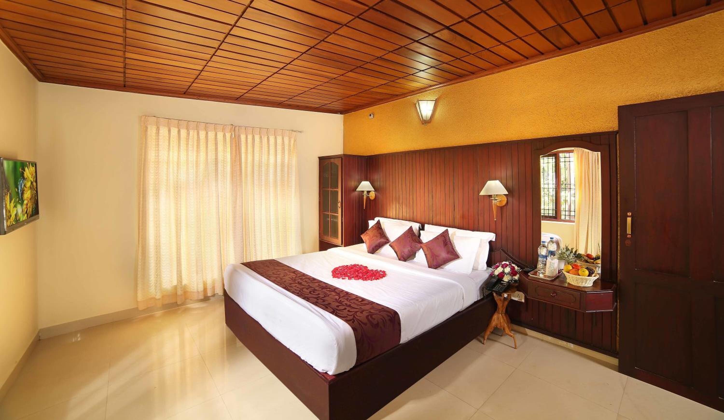 Habitación de hotel con techo de madera, una cama grande con colcha blanca y marrón, dos lámparas de pared, una mesita de noche con frutero y un armario de madera.