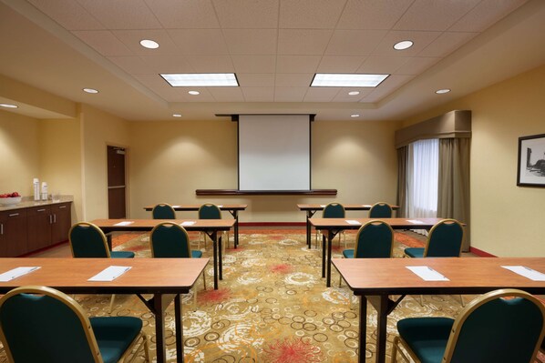 Un salón de conferencias con suelo alfombrado, una pantalla de proyector y una pizarra. Hay mesas y sillas dispuestas para reuniones.
