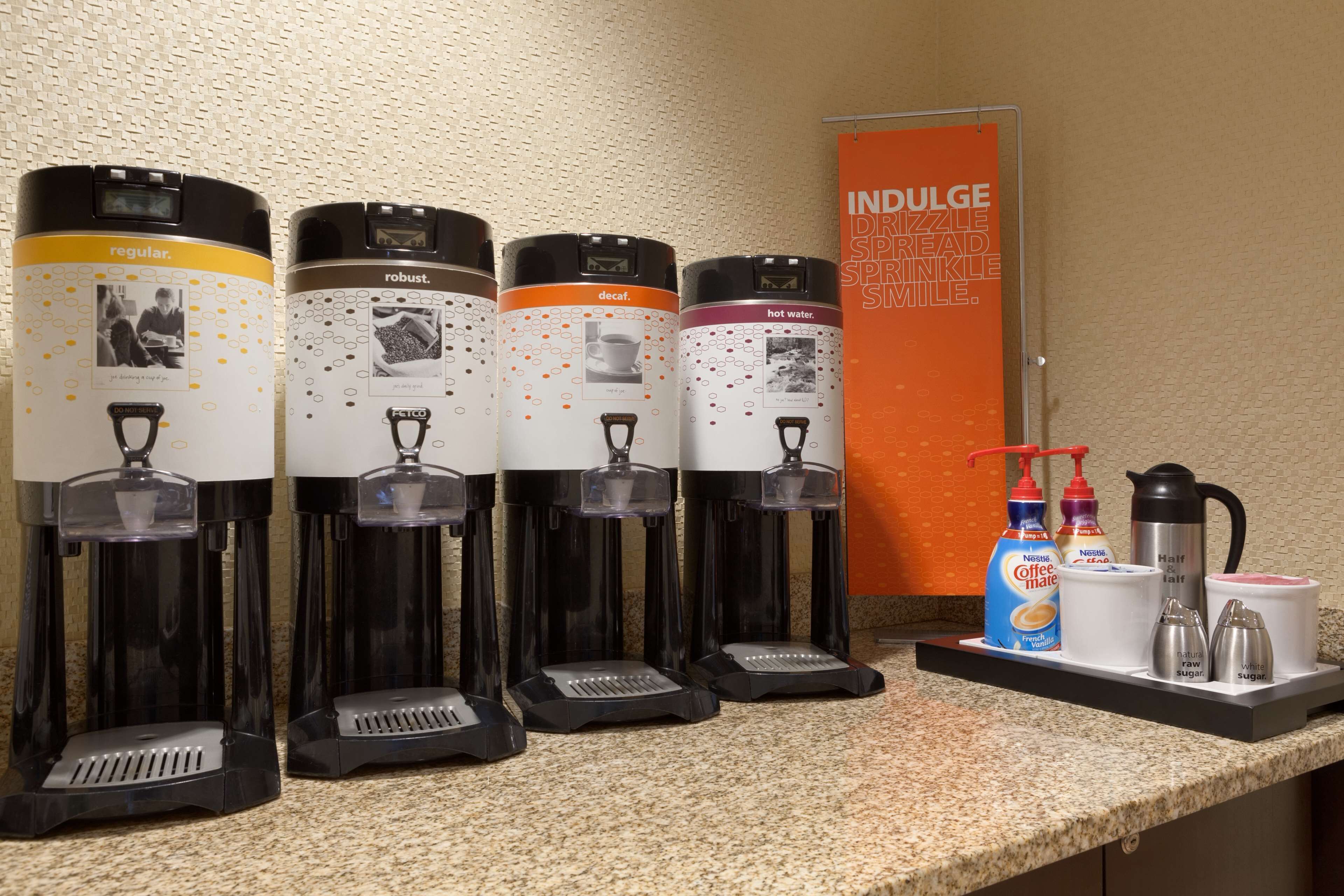 Quatre distributeurs de café étiquetés « café normal », « café corsé », « café décaféiné » et « eau chaude », ainsi qu’une machine à café, du sucre et de la crème, sont disposés sur un comptoir.
