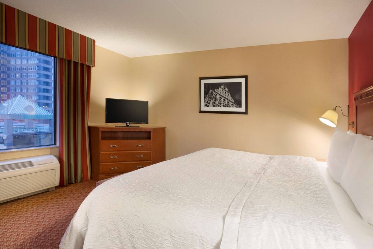 Une chambre d’hôtel avec un grand lit, un téléviseur posé sur une commode en bois, un tableau encadré au mur et une fenêtre avec des rideaux.