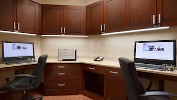 A corner office with two desks, each with a computer, a telephone, and a chair. There are wooden cabinets above the desks and a carpeted floor.