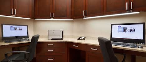 A corner office with two desks, each with a computer, a telephone, and a chair. There are wooden cabinets above the desks and a carpeted floor.