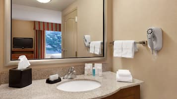 Un baño de hotel con un espejo grande, un lavabo, un secador de pelo y un televisor al fondo.