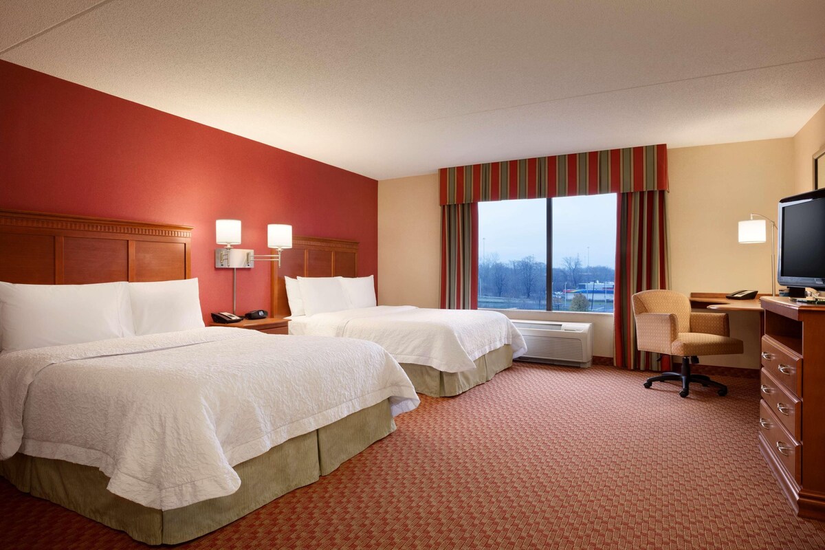 A hotel room with two beds, a desk, a chair, a television, and a window with curtains.