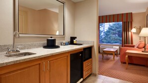 Habitación de hotel con cocina americana, que incluye un lavamanos, microondas y cafetera. Se observa una ventana con cortinas, una pequeña mesa y un sofá con una lámpara.