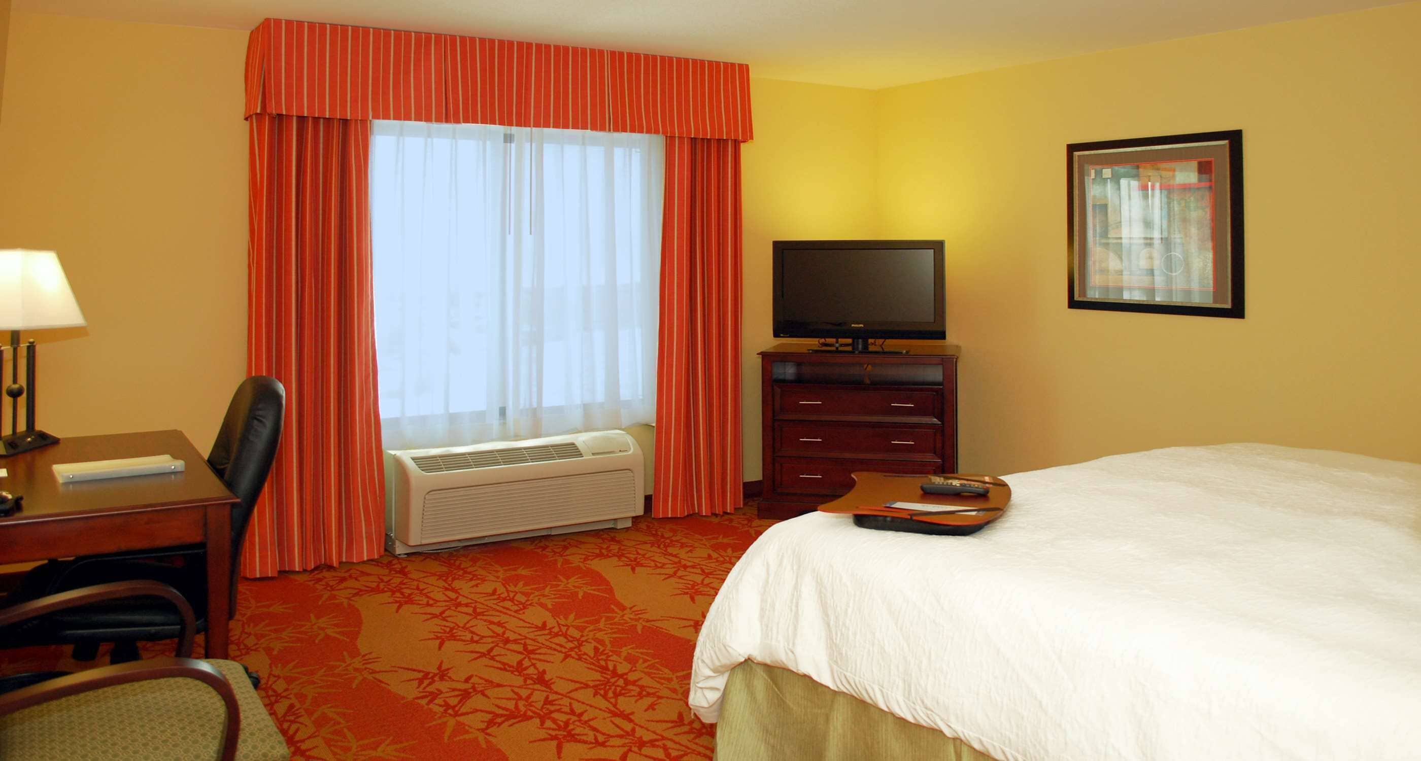 A hotel room with a bed, a desk with a lamp, a television, and a window with curtains.