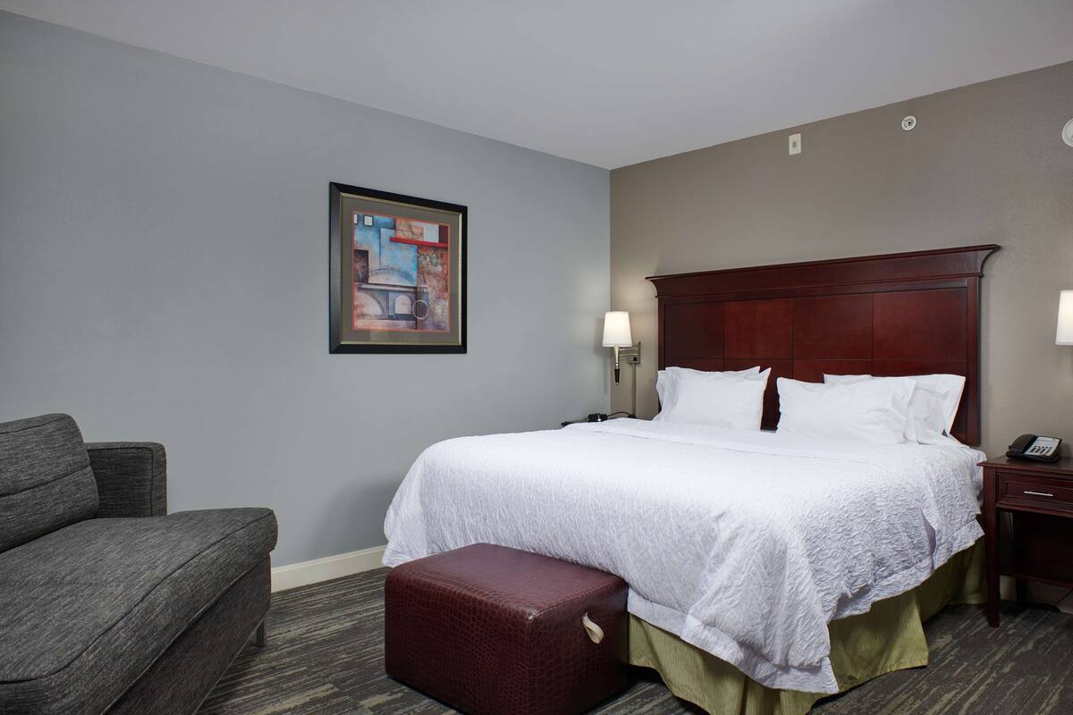 A hotel room with a large bed, a chair, a small ottoman, a nightstand with a lamp, and a framed picture on the wall.