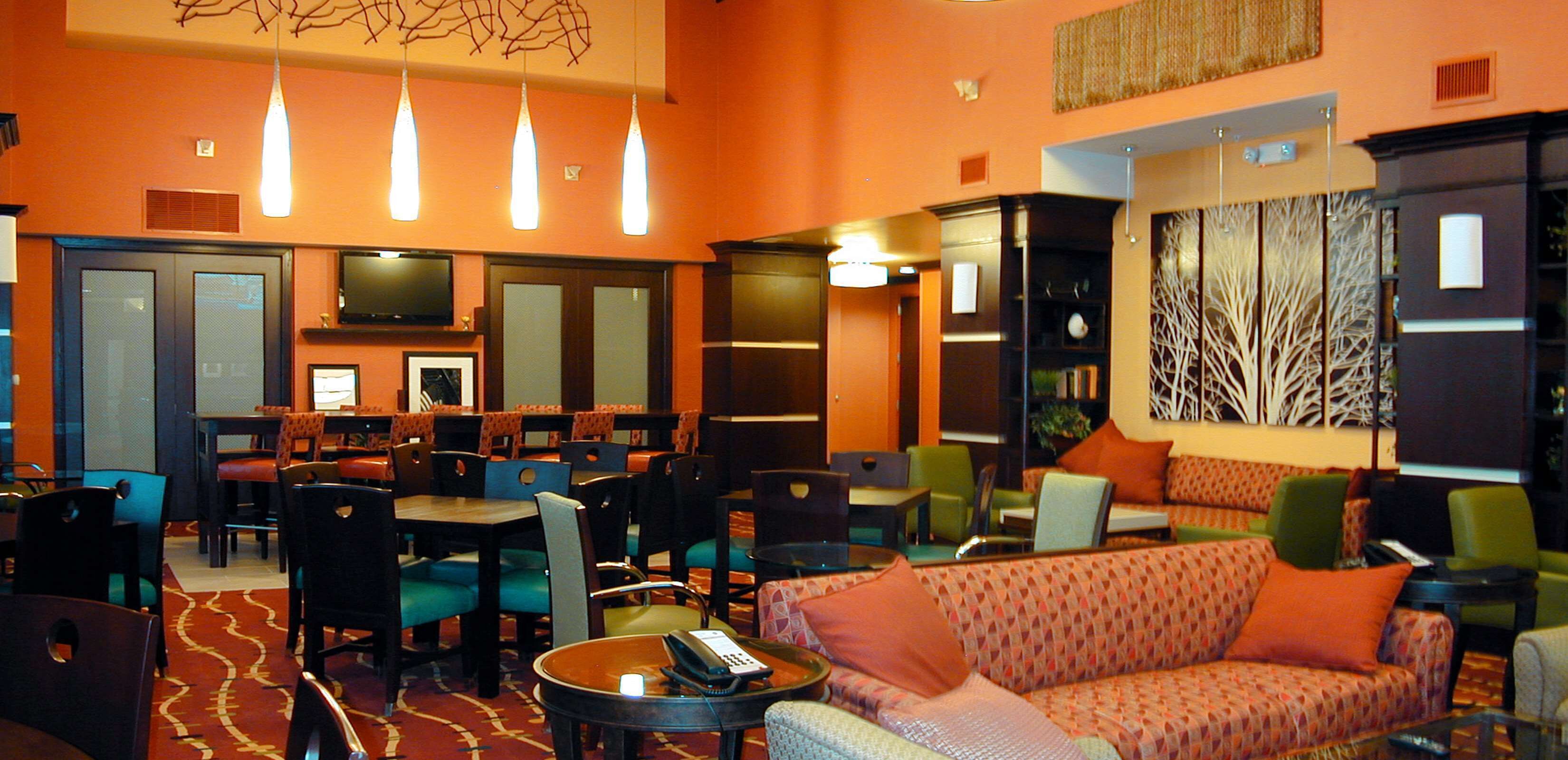 A hotel lobby with multiple seating areas, including a sofa, chairs, and tables. There are hanging lights, a television, and a decorative wall panel.